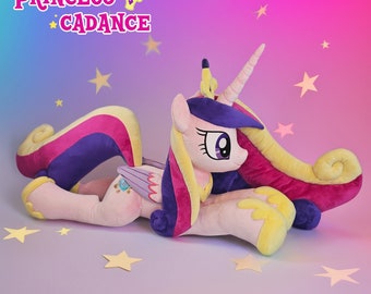 May include: A plush toy of Princess Cadance from My Little Pony. The toy is pink, yellow, and purple with a long, flowing mane and tail. The toy is lying on its side with its head turned to the right. The toy has a crown on its head and a small, blue gem on its chest.