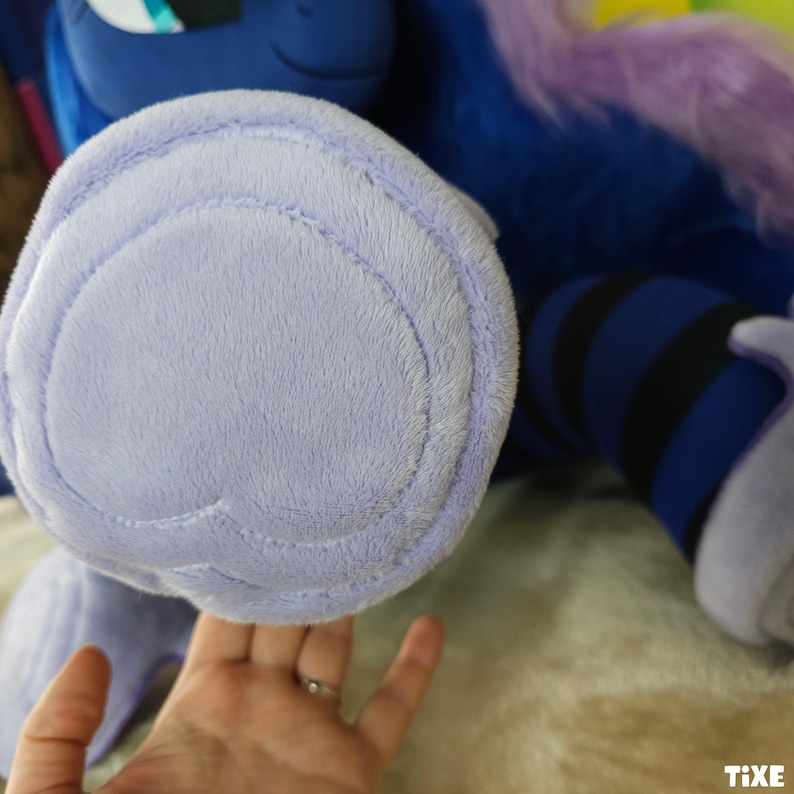 May include: Close-up of a plush toy foot in light purple. The foot is soft and plush, with a stitched outline. The toy is part of a larger character, with blue and purple elements visible in the background.