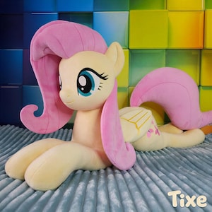 Könnte beinhalten: Ein großes Plüschtier eines gelben Ponys mit rosa Mähne und Schweif, liegend auf einer blauen Oberfläche. Das Pony hat große blaue Augen und befindet sich in einer entspannten Pose. Das Spielzeug ist weich und kuschelig, für Kinder.