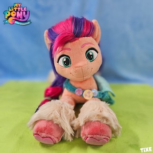 Könnte beinhalten: Ein Plüschpferd aus der Serie "My Little Pony: A New Generation". Das Pony hat einen pfirsichfarbenen Körper, rosa und lila Haare und große grüne Augen. Es trägt eine türkisfarbene Halskette mit dekorativen Knöpfen und hat flauschige, hellfarbige Füße.