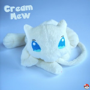 May include: Cream colored plush toy of the Pokemon Mew. The toy has blue eyes and a curled tail.