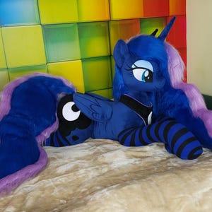 May include: A plush toy of a blue pony with a long, flowing mane and tail in shades of blue and purple. The pony has large, expressive eyes and is lying on a soft, textured surface. The pony's legs are striped with black and blue.
