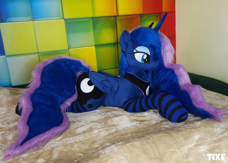 May include: A plush toy of a blue pony with a long, flowing mane and tail in shades of blue and purple. The pony has large, expressive eyes and is lying on a soft, textured surface. The pony's legs are striped with black and blue.