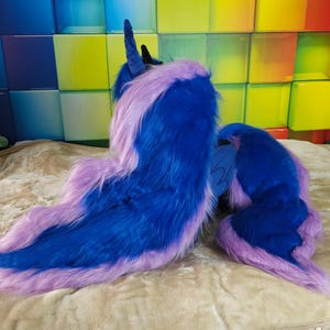 May include: A plush, blue and purple costume piece, possibly a tail or mane, with a long, flowing design. The item is made of soft, faux fur material. The colors are vibrant and the overall aesthetic is fantasy-themed.