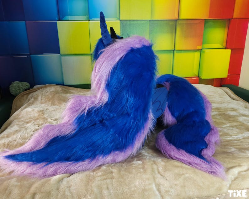May include: A plush, blue and purple costume piece, possibly a tail or mane, with a long, flowing design. The item is made of soft, faux fur material. The colors are vibrant and the overall aesthetic is fantasy-themed.