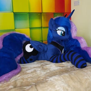 May include: A plush toy of Princess Luna from My Little Pony. The toy is primarily blue with a purple mane and tail. It has a black and white crescent moon symbol on its flank and black and blue striped legs.