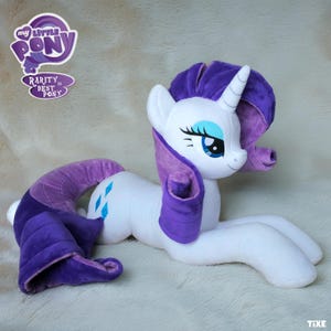 Könnte beinhalten: Ein weißes Plüschpferd mit lila Mähne und Schweif, blauen Augen und einem Einhornhorn. Das Pony liegt auf einer weichen, weißen Oberfläche. Der Text "My Little Pony" und "Rarity is Best Pony" sind sichtbar.