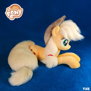 May include: A plush toy of the character Applejack from My Little Pony. The toy is yellow with a cream-colored mane and tail. It has green eyes and a red ribbon around its forelock. The My Little Pony logo is in the upper left corner.