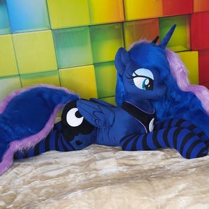 May include: A plush toy of Princess Luna from My Little Pony. The toy is primarily dark blue with a purple mane and tail. It has black and blue striped legs and is lying on a beige surface. The background features a colorful, geometric pattern.