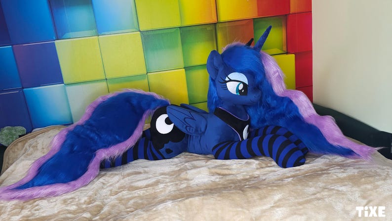 May include: A plush toy of Princess Luna from My Little Pony. The toy is primarily dark blue with a purple mane and tail. It has black and blue striped legs and is lying on a beige surface. The background features a colorful, geometric pattern.