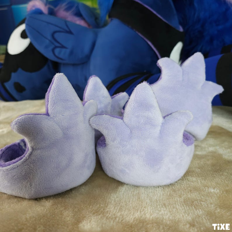 May include: Close-up of three light purple plush paw pads with a three-pointed design. The paw pads are made of soft, fuzzy material and have a darker purple interior. A blue plush toy is in the background.