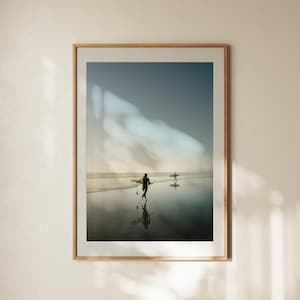 May include: Framed print of a beach scene with two surfers walking towards the ocean. The image has a muted color palette with a light blue sky and a sandy beach. The frame is made of light-colored wood.