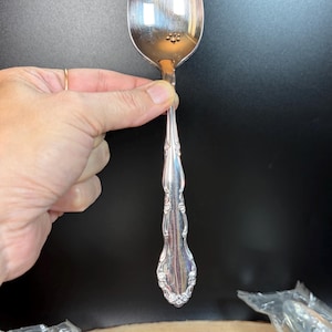 May include: A silver spoon with an ornate handle. The spoon has a rounded bowl and a long, detailed handle with floral and scroll designs. The spoon is held against a black background, with a wooden surface visible at the bottom.