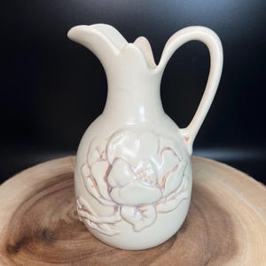 May include: Off-white ceramic pitcher with a floral design. The pitcher has a handle and a spout. The floral design is embossed on the front of the pitcher. The pitcher is sitting on a wooden surface.