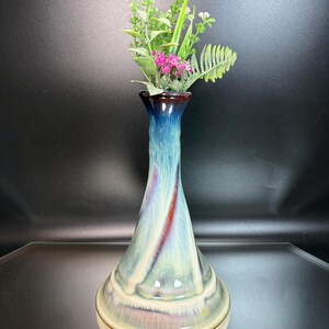 May include: A tall, decorative vase with a flared base and a slender neck. The vase features a gradient of colors, including blue, green, and brown, with a swirl of red. It is filled with a small arrangement of artificial flowers and greenery.