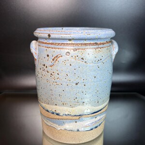 May include: A cylindrical ceramic vase featuring a light blue speckled glaze with dark brown flecks. The vessel has two small side handles, a thick rim, and a lower section with horizontal bands of beige, blue, and dark blue layered glaze.