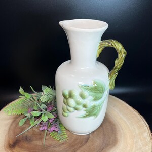 May include: A white ceramic jug featuring a green grape and leaf design. The handle is a twisted green vine. The jug is displayed on a wooden surface with greenery.
