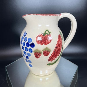 May include: A white ceramic pitcher with a red rim and a handle. The pitcher is decorated with hand-painted fruit designs, including grapes, strawberries, and a red pepper.