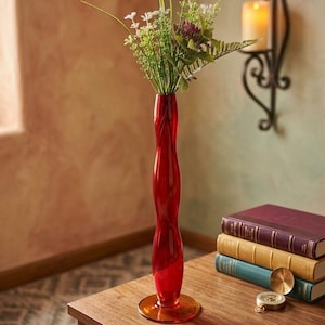 Puede incluir: Un florero de vidrio rojo, alto y esbelto, con un cuerpo ondulado y una base circular ancha. El florero contiene un ramo de follaje verde, flores blancas y una pequeña flor morada. El vidrio es translúcido, permitiendo el paso de la luz.