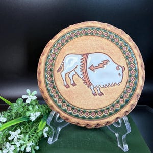 Puede incluir: Plato redondo de cerámica con fondo beige y borde decorativo. El centro presenta un bisonte blanco con un diseño de rayo. El borde tiene un patrón geométrico verde y marrón.