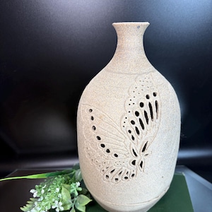 Puede incluir: Un jarrón de cerámica beige con un diseño de mariposa tallado en la superficie. Las alas de la mariposa están parcialmente recortadas, creando un patrón calado. El jarrón tiene un cuerpo redondeado y un cuello estrecho.