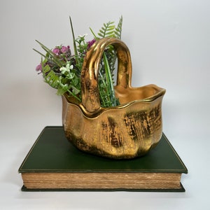 May include: A gold-colored ceramic planter shaped like a basket with a handle. The planter is sitting on a green book and has a bouquet of purple and white flowers inside.
