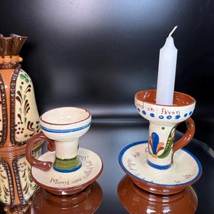 May include: Two ceramic candle holders with handles and a tall vase. The holders feature cream, brown, blue, and green hand-painted floral patterns and text. One holder contains a white taper candle and is inscribed with Stratford-on-Avon.