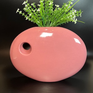 May include: A pink ceramic vase with a unique oval shape and a small hole on the side. The vase is filled with a bouquet of white flowers and green leaves.