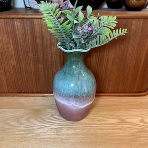 Op de afbeelding: Een groene en bruine keramische vaas met een gegolfde rand, gevuld met een boeket groene varens en paarse bloemen.