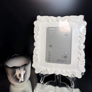 May include: A white, ornate picture frame with a decorative floral design. The frame is labeled ".KVILL" and is designed to hold a 5 x 7 inch photo. A black and white decorative vase is to the left of the frame.