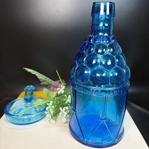 May include: A vibrant blue glass bottle with a unique bubble-like design on the upper portion. The bottle has a matching lid and is decorated with geometric patterns. The text "WEAVER'S AMBER" is visible on the bottle. Decorative flowers are next to the bottle.