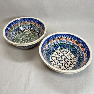 May include: Two round ceramic bowls featuring a detailed blue, green, and orange floral pattern. The interior of each bowl is decorated with a repeating geometric design of small orange dots and blue swirls, finished with a blue rim and white base.