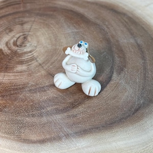 May include: A small, white ceramic dog figurine with brown ears and a bone in its mouth. The dog has blue eyes and is sitting with its paws clasped. The figurine is on a wooden surface.
