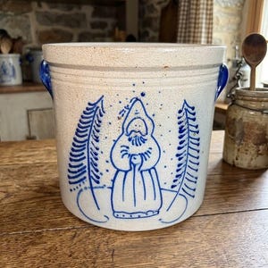 May include: A cylindrical stoneware crock with a speckled off-white glaze and blue hand-painted details. The design features a central figure of a bearded person in a hooded robe flanked by two stylized evergreen trees and blue side handles.