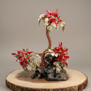 May include: A decorative sculpture featuring three clusters of red and cream-colored seashell flowers with small green beads at their centers. The flowers are attached to a central copper-colored wire stem anchored to a dark, textured piece of driftwood.