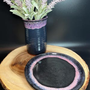 May include: A handcrafted ceramic plate with a black centre and a pink and purple rim rests on a wooden slab. A dark blue vase with a purple rim holds lavender stems. The vase and plate are on a dark background.