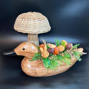 Könnte beinhalten: Eine hölzerne Entenfigur mit einem geflochtenen Korb-Pilz-Hut. Die Ente ist mit verschiedenen Nüssen und Grünzeug verziert.