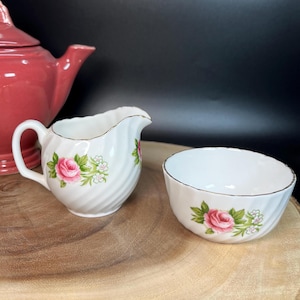 Op de afbeelding: Een witte roomkan en suikerpot set met een roze roos en groen blad ontwerp. De set heeft een gouden rand en een swirl patroon.
