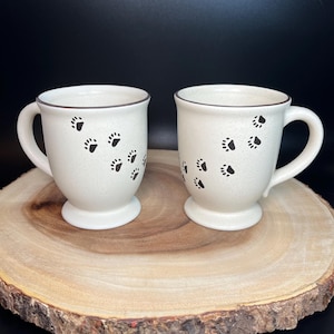 May include: Two white ceramic mugs with brown rims and black paw print designs on a wooden surface.