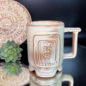 Op de afbeelding: Een beige keramische mok met een bruin reliëfontwerp met een medisch symbool en het woord "AECCPOMS". De mok heeft een vierkant handvat en wordt weerspiegeld op een glanzend oppervlak. Een decoratieve plaat en een vetplant staan op de achtergrond.
