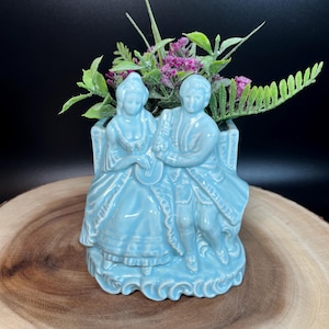 Könnte beinhalten: Hellblaue Keramikvase mit einem sitzenden Paar in historischer Kleidung. Die Vase ist mit künstlichem Grün und lila Blumen gefüllt. Die Vase hat einen dekorativen Sockel und steht auf einer Holzoberfläche.