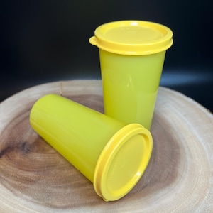 May include: Two lime green plastic tumblers with yellow lids. One tumbler is upright, and the other is lying on its side. The tumblers are on a wooden surface with a dark background.