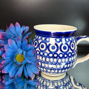 Puede incluir: Una taza de cerámica redonda con base blanca y un patrón repetitivo de círculos y puntos de color azul oscuro. La taza tiene un asa gruesa y un borde ancho. Junto a la taza hay varias flores artificiales de color azul brillante con centros amarillos.