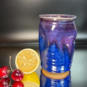 May include: A cylindrical ceramic tumbler featuring a glossy glaze that transitions from deep royal blue at the base to a rich purple at the rim. The surface has a textured, dimpled appearance. Two red cherries and a lemon slice sit nearby.
