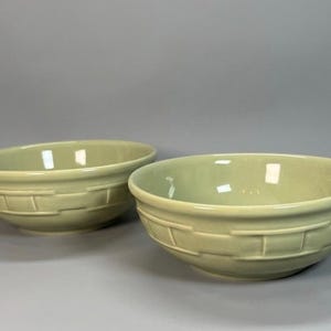 May include: Two matching round bowls in a pale sage green colour. Each bowl features a raised, embossed basket-weave pattern around the exterior side. The interior of the bowls is smooth and glossy, with a wide opening and a rounded base.