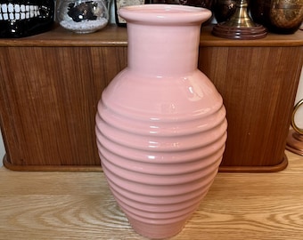 May include: A pink ceramic vase with a ribbed design. The vase is sitting on a wooden surface.
