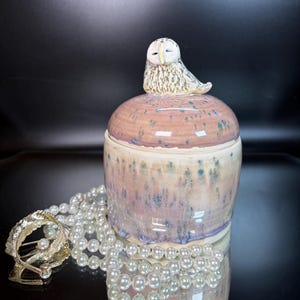 May include: A ceramic jar with a lid topped by a sculpted owl figurine. The jar features a glazed finish in shades of pink, cream, and blue with vertical drip patterns. Beside the jar lies a gold-toned leaf-shaped brooch and a strand of white pearls.