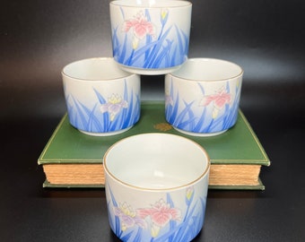May include: Set of four white ceramic teacups with a blue and pink floral design. The cups are stacked on top of each other and are sitting on a green book.