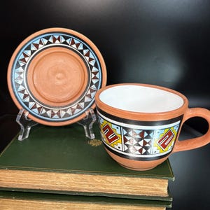 May include: A terracotta-colored ceramic teacup and saucer set. The cup and saucer feature geometric patterns in black, white, blue, and yellow. The cup has a handle and a white interior. The saucer is round with a raised center.