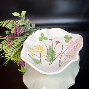 May include: A white, scalloped-edge ceramic bowl with a floral design featuring green clovers, yellow and pink flowers. The bowl is resting on a light green base, with a sprig of purple flowers and green leaves to the left.
